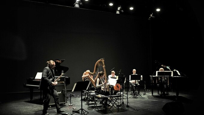 A group of musicians performing on stage including a harpist, pianist, bassist, and other musicians.