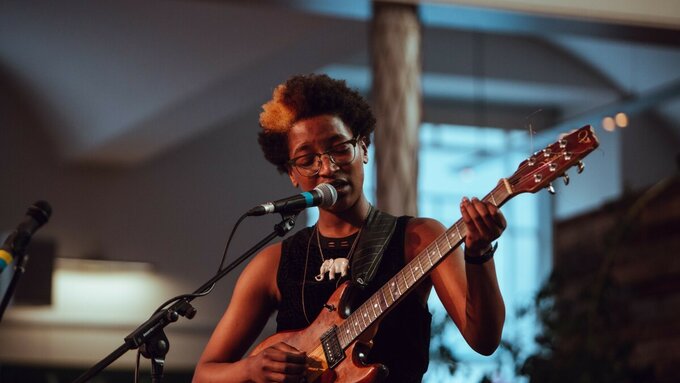 A musician wearing glasses sings into a microphone whilst playing the guitar.