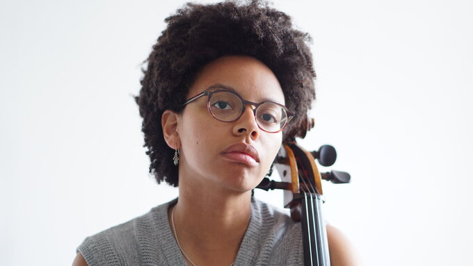 A performer wearing glasses, and a grey jumper sits with a cello in front of a white background.