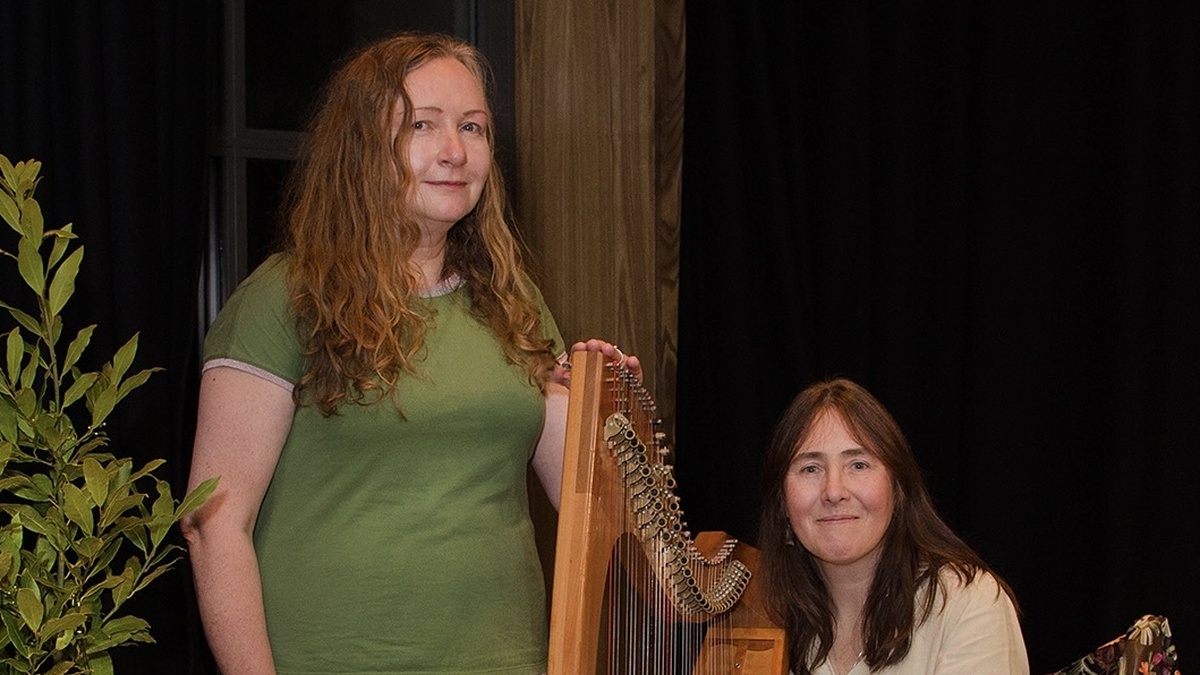 Two people, one standing and one sitting with a harp in a room with plants.