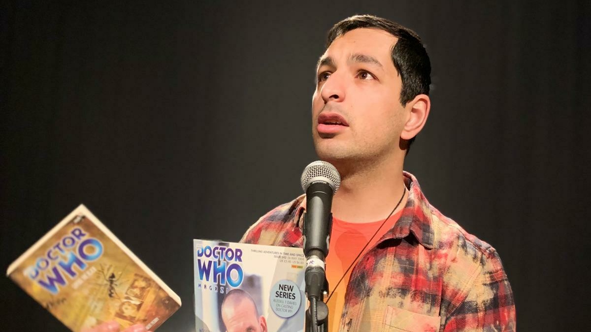 A person wearing an orange t-shirt and checked shirt talks into a microphone, holding two Dr Who books.