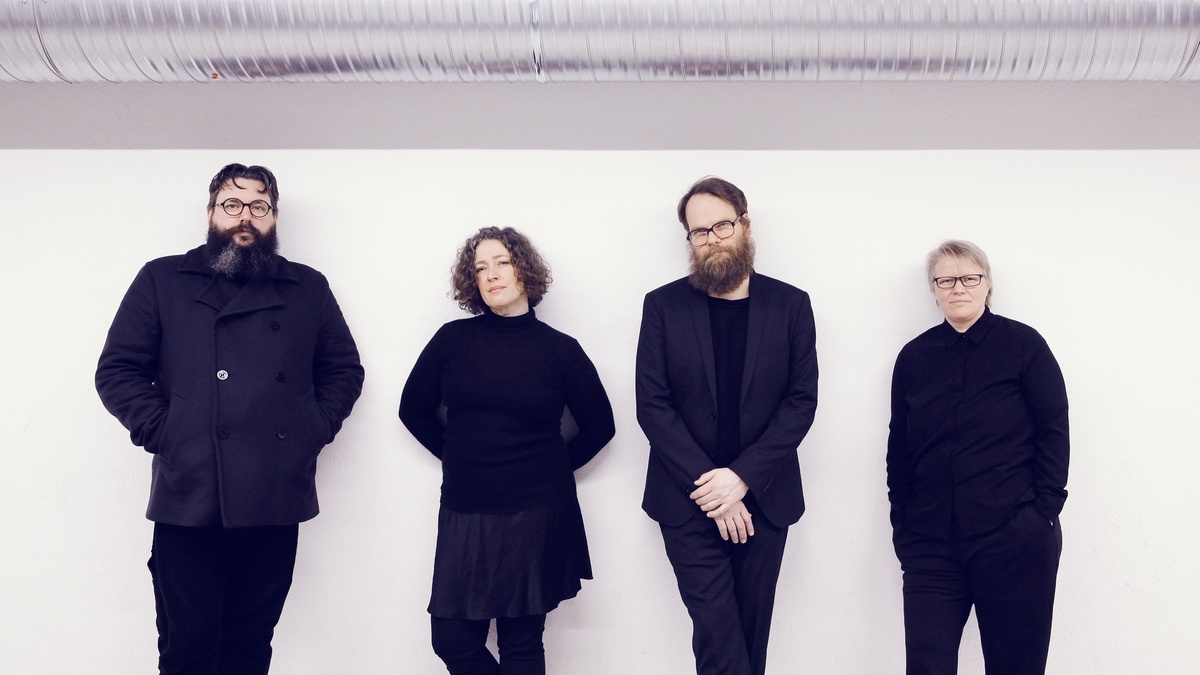 Four people wearing all black leaning against a white wall.