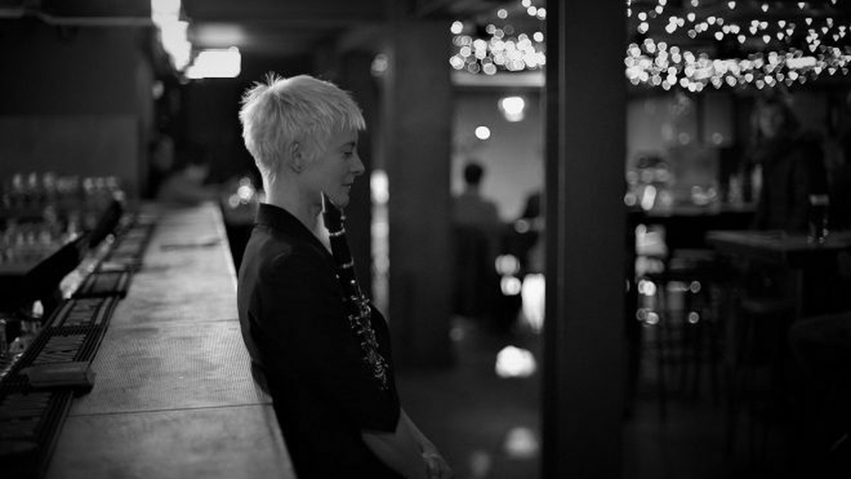 A black and white androgynous person with short bleached hair leaning against a bar holding a clarinet.