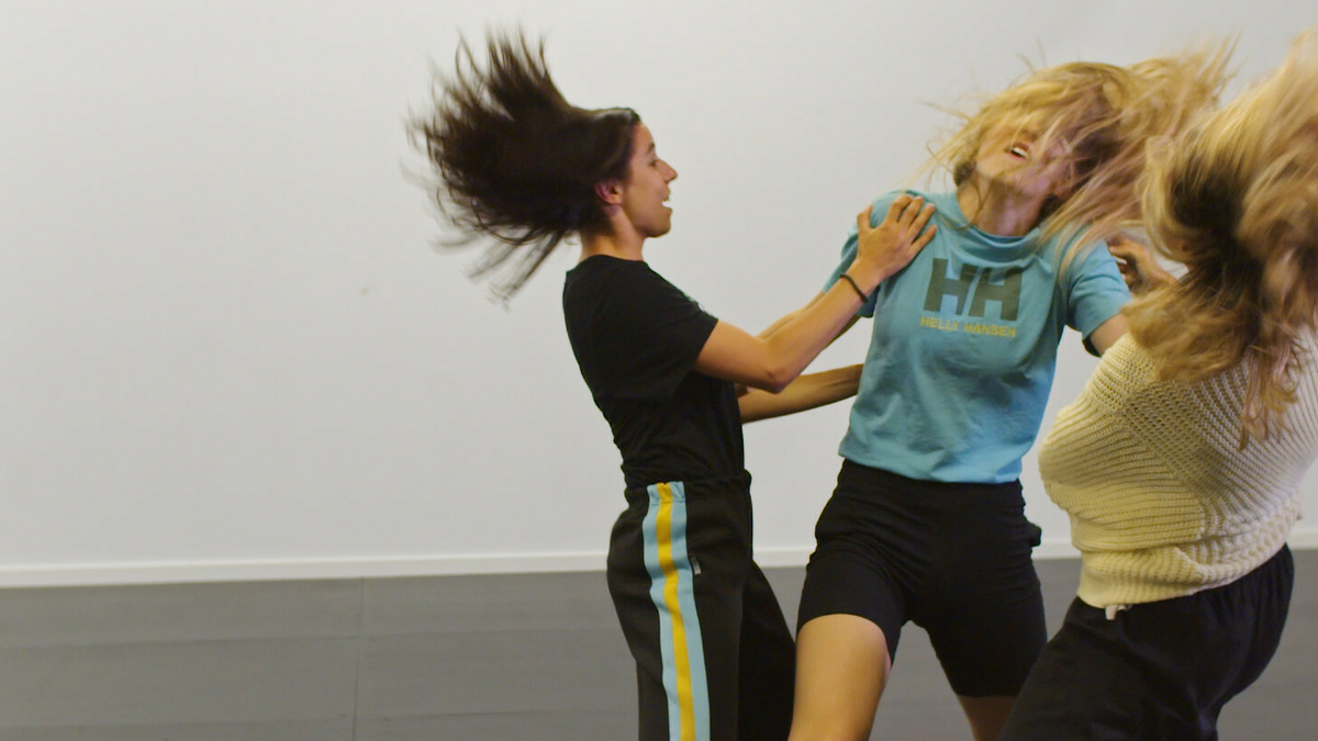 Three people dancing together in a dance studio with white walls and wooden flooring.