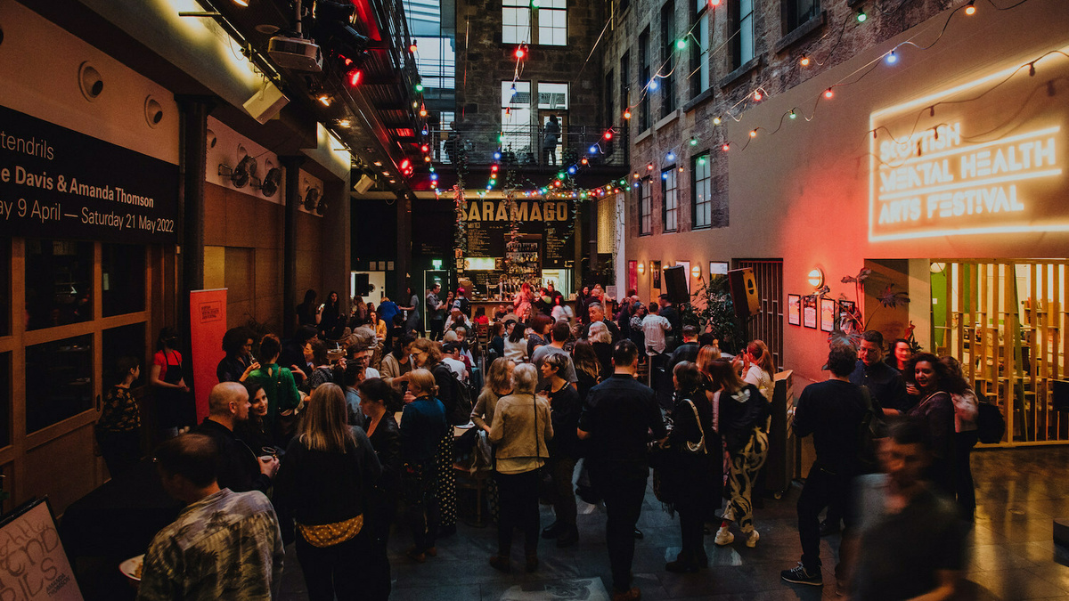 A busy room full of people chatting and having some drinks with colourful fairy lights overhead.