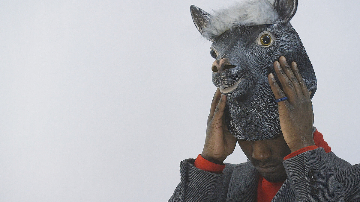 A person wearing a red jumper and grey jacket takes off a grey lama mask in front of a white background.