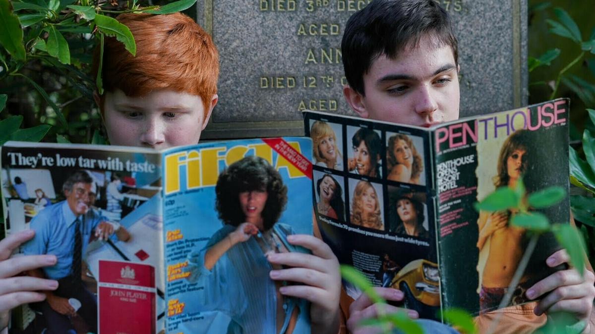 Two young people sit in front of a gravestone reading magazines.