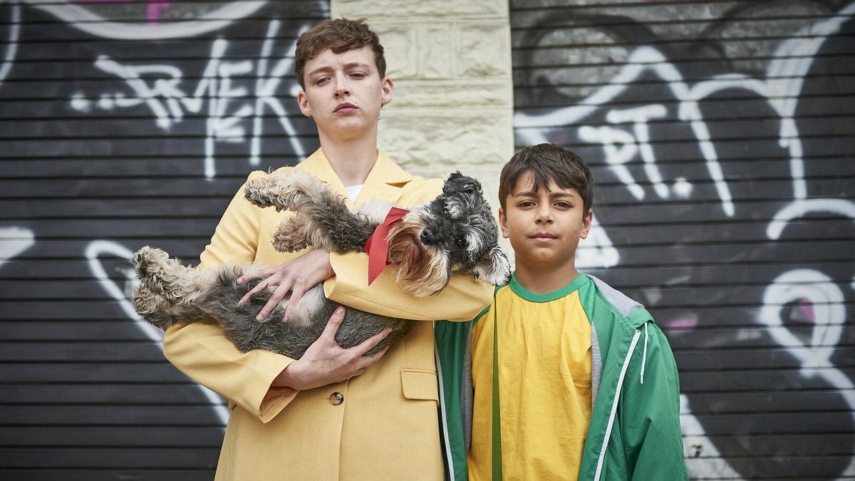 Two boys stand in front of a graffitied backdrop. One is cradling a dog in his arms.