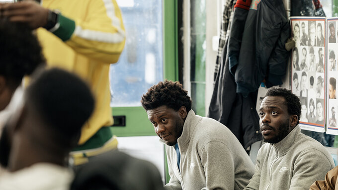 Two men sit in a barber's shop. They look up at the person getting their hair cut.