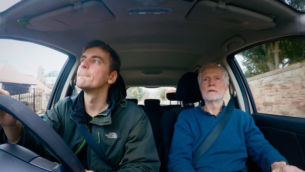 Two men sit in a car - the younger one is driving, the older one is in the passenger seat.