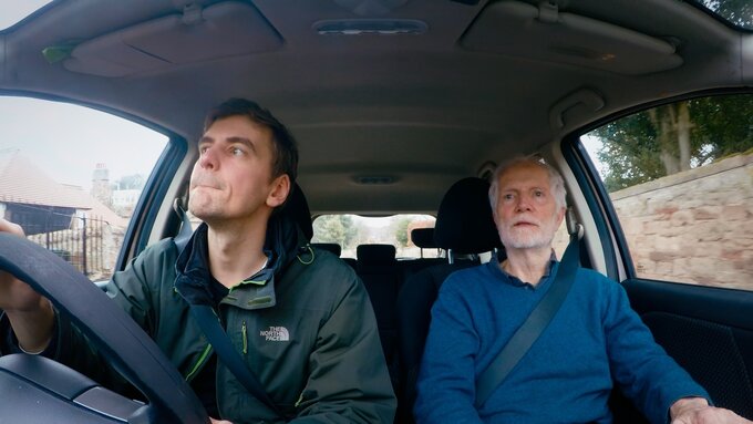 Two men sit in a car - the younger one is driving, the older one is in the passenger seat.