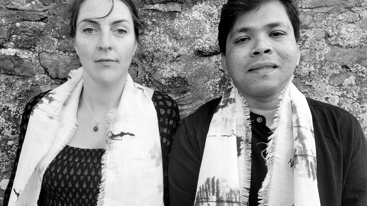 A black and white picture shows a man and woman each wrapped in a scarf standing in front of a large stone wall.