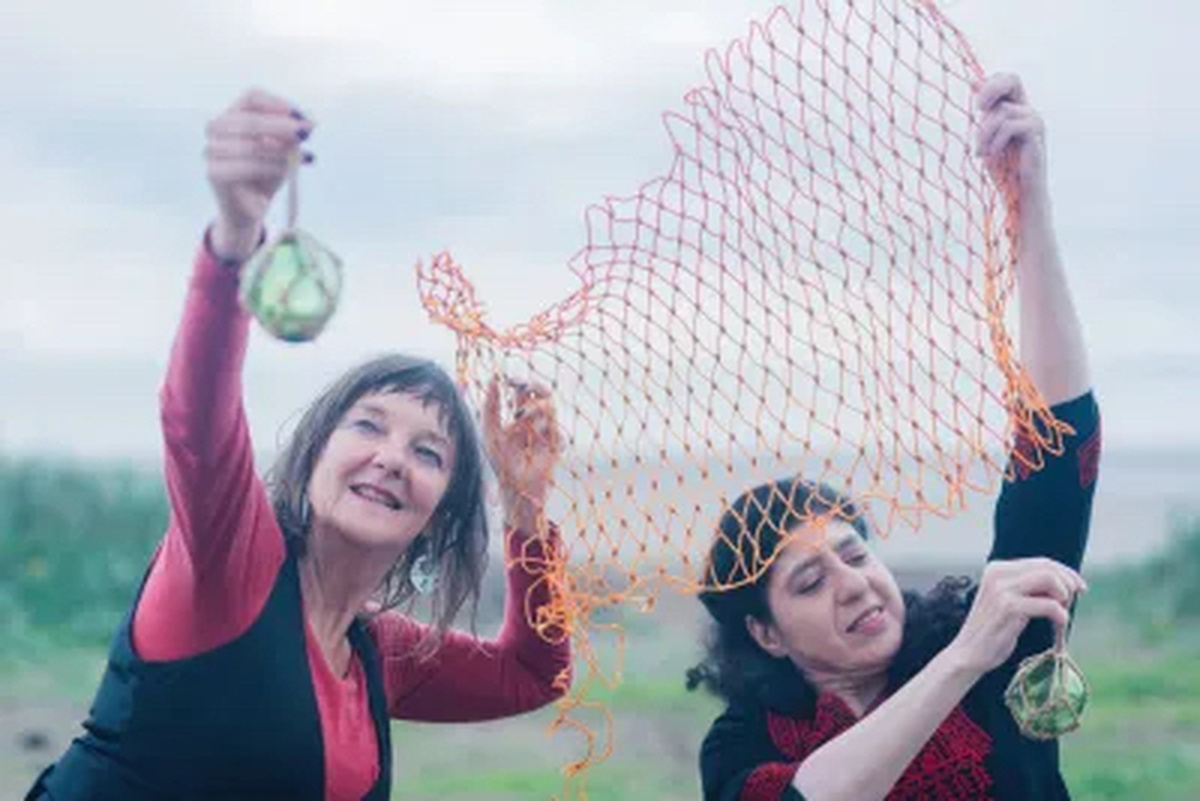 Two women play with a piece of orange fishing net and they both suspend green glass baubles from their hands.