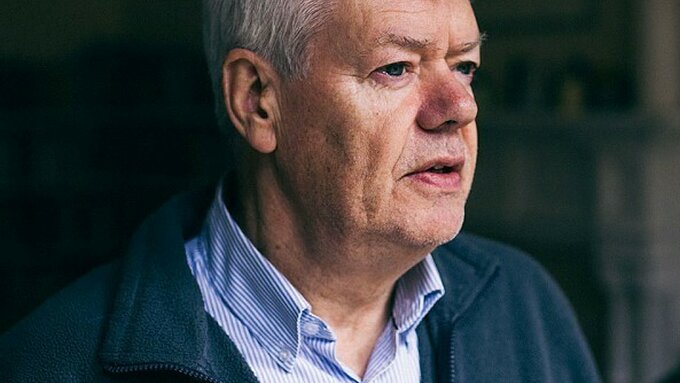 An older man wearing a blue pinstripe shirt and fleece.