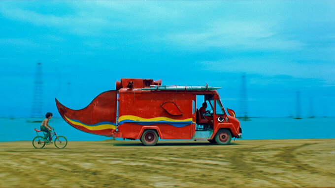 A brightly coloured van with a tail travels along a landscape with a blue sky, with a child on a bicycle following