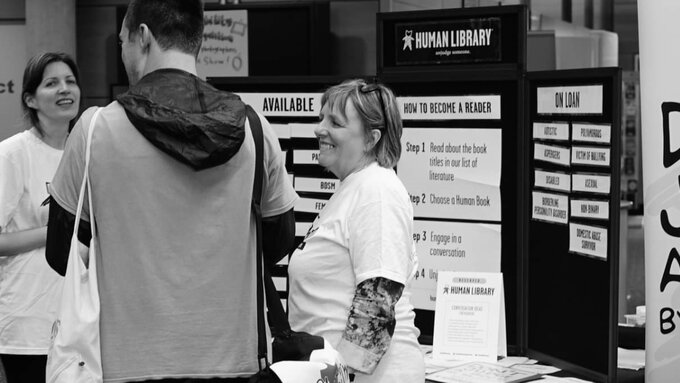 A black and white photo of people gathered at a 'human library' stall. A banner reads "Don't judge a book by its cover".