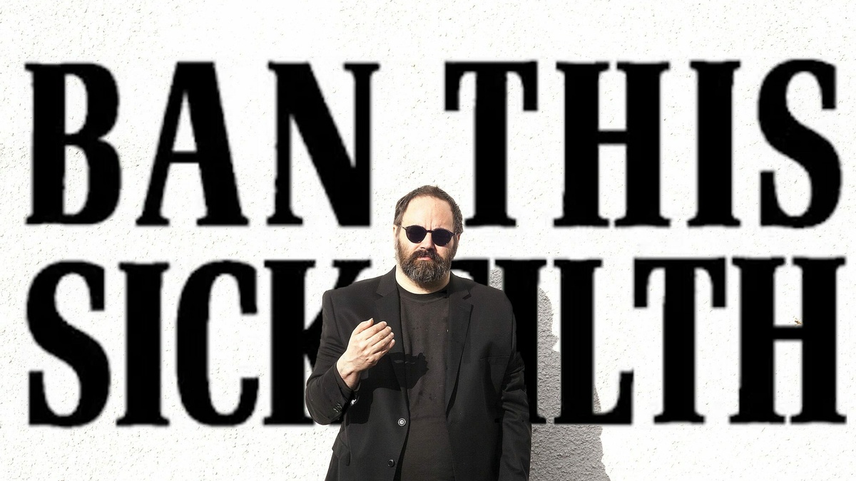 A man wearing black clothes and sunglasses stands in front of a sign that says 'Ban this sick filth'.