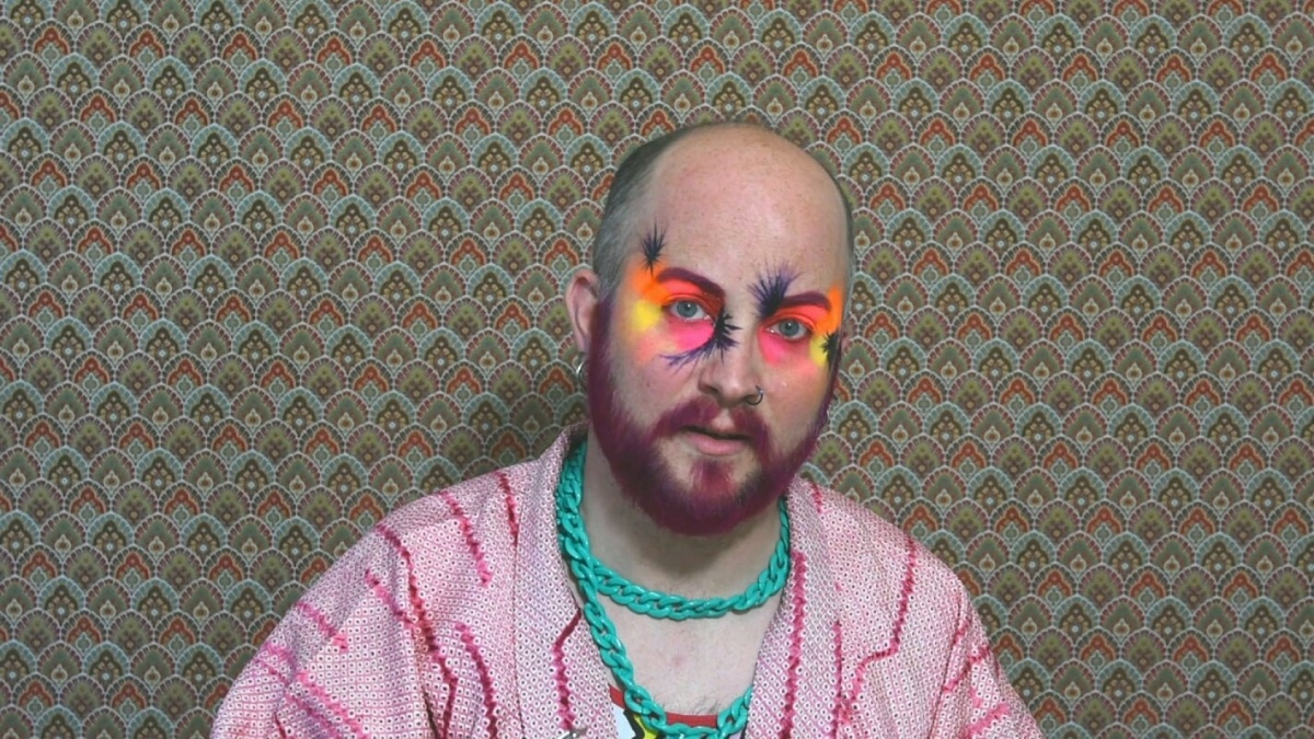 A man looks straight at us. He has a pink beard and colourful eye makeup. He's standing against a patterned wallpaper.