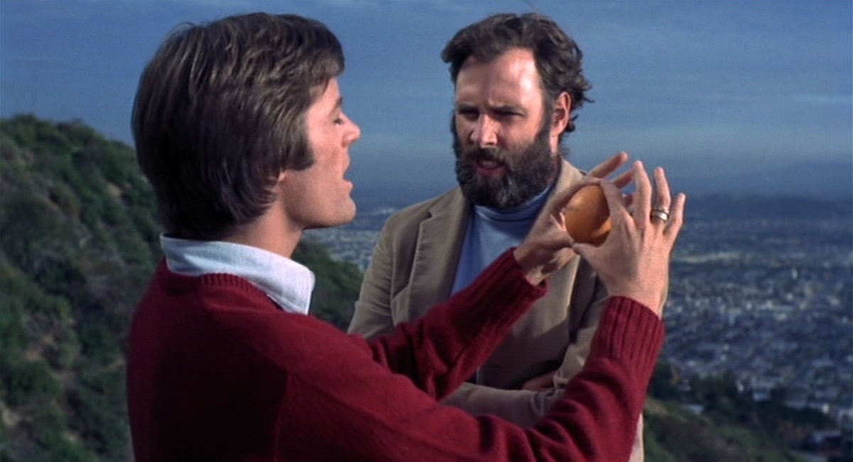 Two men talk on a cliff face, one of them is holding out an orange