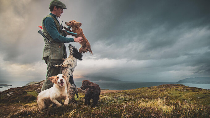 A gamekeeper surrounded by dogs stands on a mountainside overlooking the sea.