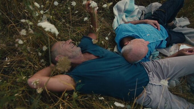 Two men lie in a field surrounded by flowers. One man is resting on the other, they both look relaxed and happy.