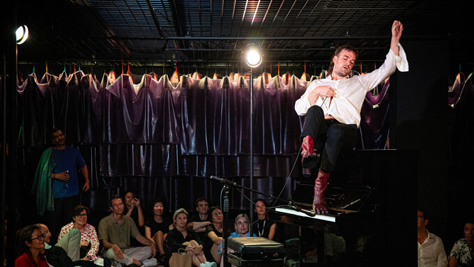 A man reclines on a large piano holding a microphone, the absurd red fingered tabi boots he wears rest on the keys.