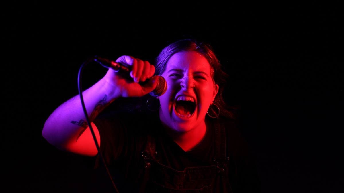 A young femme with hoop earrings looking toward the camera, aggressively screaming into a microphone.