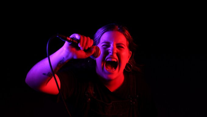 A young femme with hoop earrings looking toward the camera, aggressively screaming into a microphone.