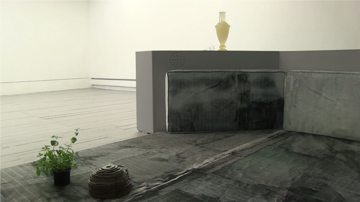 A gallery space has a large grey structure placed across the floor, with a potted plant a small sculpture on top.