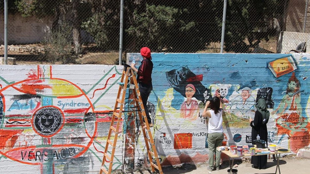 Members of the International Academy of Art Palestine paint a wall in Palestine with scenes of people in bright colours.